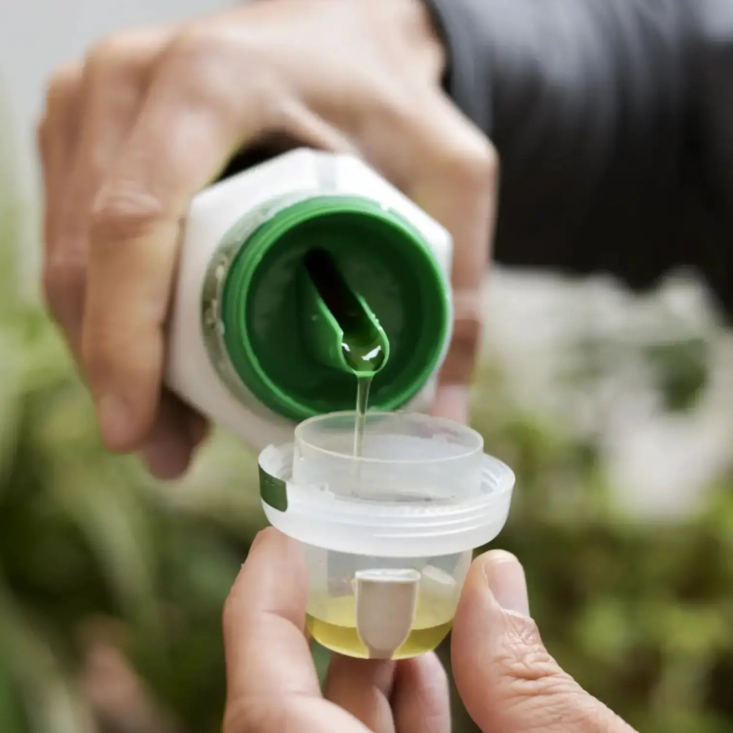 Person gießt Flüssigkeit aus einer grün-weißen Flasche in einen kleinen Behälter vor unscharfem natürlichem Hintergrund